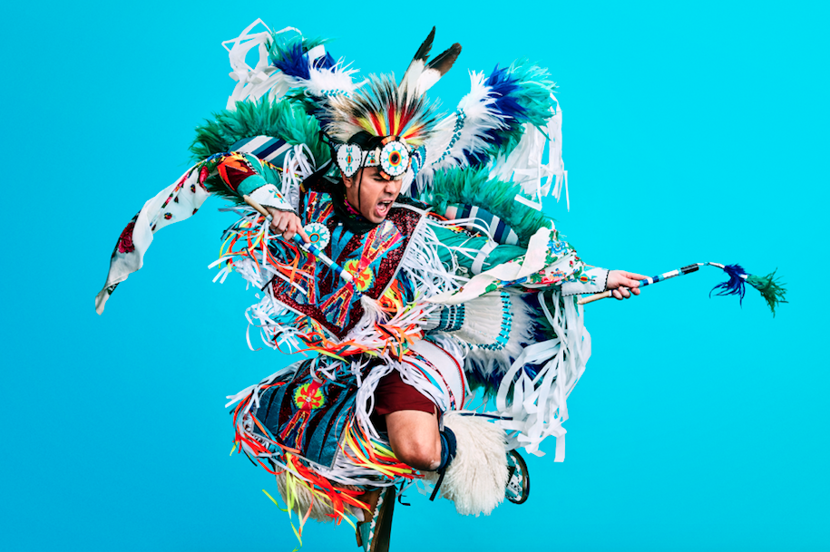 Notorious Cree in traditional Indigenous regalia jumping up against blue background